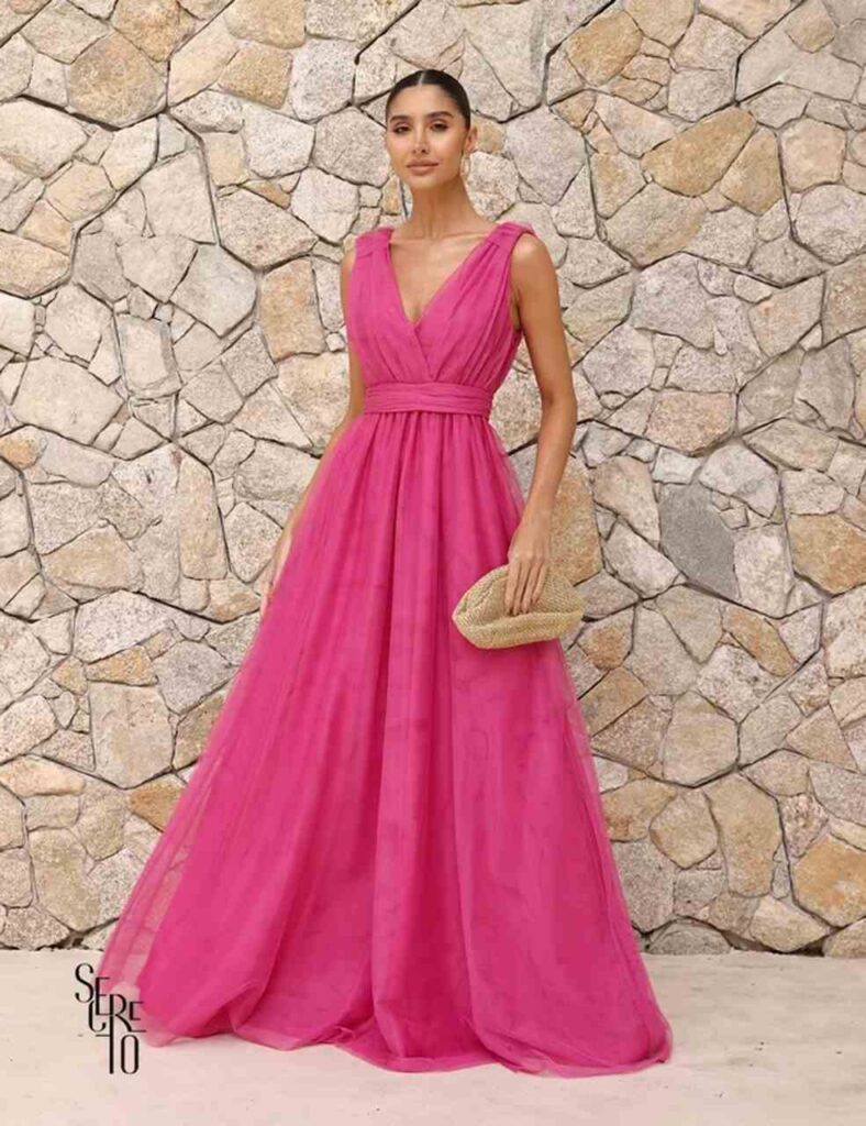 Mulher posa com vestido para baixinhas: longo fúcsia, decote profundo, cintura marcada e tecido fluido que valoriza a silhueta com elegância