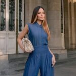 Pessoa em frente a colunas clássicas, vestindo conjunto jeans azul moderno com bolsa clara elegante, destacando estilos de roupas casuais sofisticados.