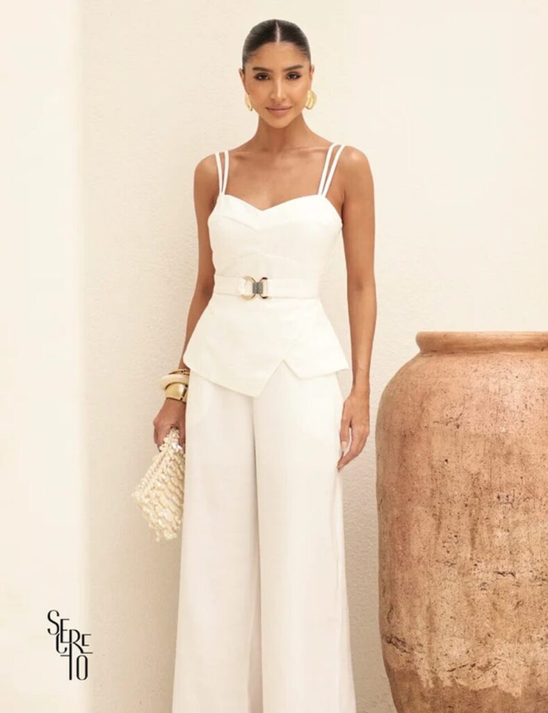 Mulher elegante com look de ano novo: conjunto branco com top de alças duplas e calça ampla, acessórios dourados, clutch com pérolas, penteado preso, em ambiente minimalista com vaso rústico.