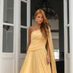 Pessoa em vestido amarelo longo e elegante, modelo de um ombro só com drapeado sofisticado, cenário luxuoso com portas brancas e lustre, inspiração perfeita para looks-pré-wedding glamorosos.