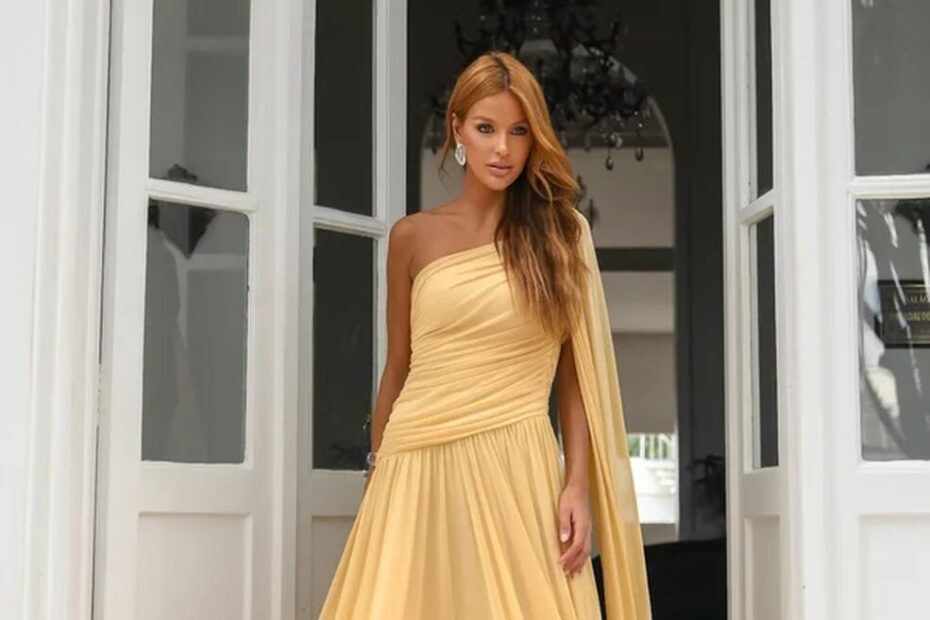Pessoa em vestido amarelo longo e elegante, modelo de um ombro só com drapeado sofisticado, cenário luxuoso com portas brancas e lustre, inspiração perfeita para looks-pré-wedding glamorosos.