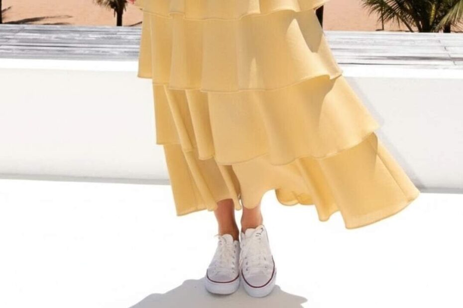 Pessoa em praia tropical usando vestido amarelo com babados e tênis branco, exemplo charmoso de looks vestido longo com tênis para estilo casual elegante.
