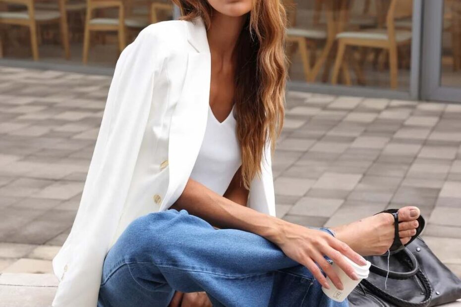 Mulher sentada em banco de concreto, usando blazer com calça jeans, óculos escuros e segurando xícara, estilo editorial moderno urbano.